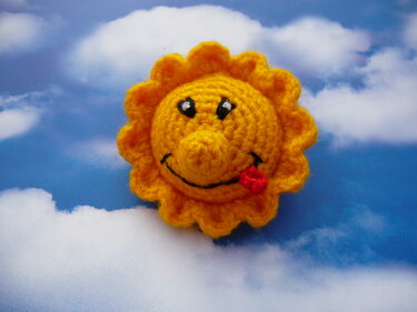 Kleines gehäkeltes Sonnen-Amigurumi mit lachendem Gesicht und roter Zunge auf wolkigem Hintergrund.