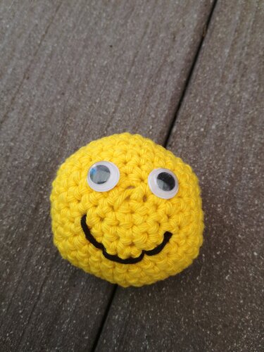 Gelber, gehäkelter Smiley mit Wackelaugen und gesticktem Mund auf Holzoberfläche.