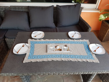 Gehäkelter Tischläufer in Beige und Hellblau auf grauem Gartentisch mit Tellern und Dekotablett.