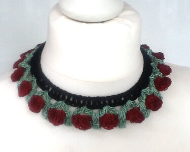 Gehäkeltes Halsband in Schwarz mit roten Beeren und grünen Schlaufen auf weißer Büste