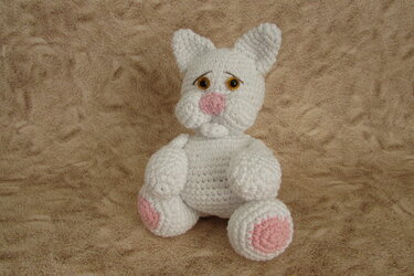 Weißes gehäkeltes Amigurumi-Kätzchen mit rosa Nase und rosa Pfotenpads sitzt auf beigem Stoff
