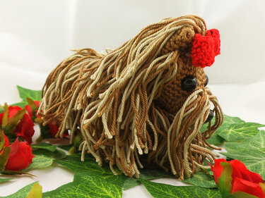 Gehäkelter Amigurumi-Hund mit zotteligem braun-beigem Garn und roter Schleife auf grünen Blättern und roten Rosen