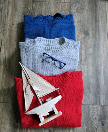 Drei gefaltete Strickpullover in Blau, Grau und Rot mit blauer Brille und Holzsegelboot auf Holzboden.