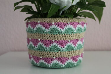 Gehäkelte Blumentopf‑Hülle mit Zickzackmuster in Rosa, Mint und Weiß, goldglänzende Reihen und aufgenähte Perlen