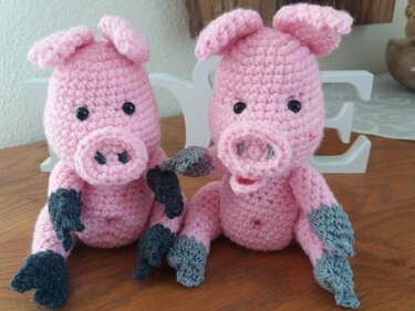 Zwei rosa gehäkelte Amigurumi‑Schweinchen mit grauen Füßen und schwarzen Augen auf Holz
