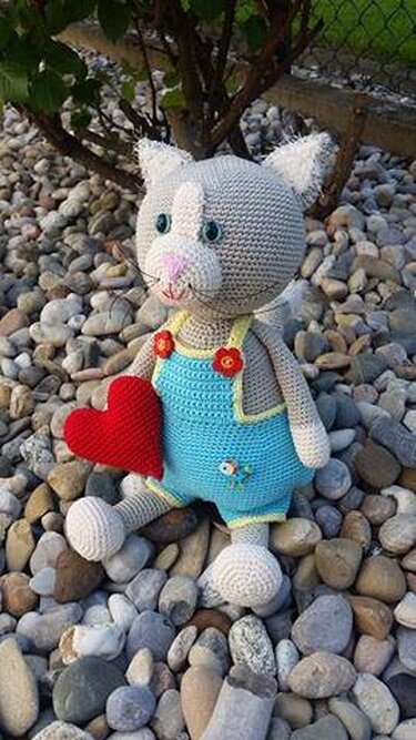 Gehäkelte graue Amigurumi-Katze im hellblauen Overall mit rotem Herz auf Kieselsteinen