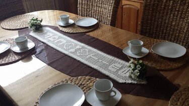 Weißer gehäkelter Tischläufer im Filetmuster auf dunklem Unterläufer, arrangiert auf einem Holztisch mit geflochtenen Platzsets und weißem Geschirr.