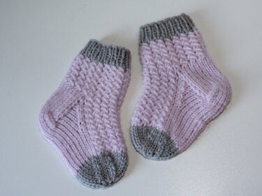 Paar gestrickter Babysocken in Flieder mit grauem Bündchen, Ferse und Spitze auf hellem Untergrund