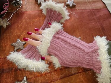 Rosa gestrickte fingerlose Armstulpen mit weißem flauschigem Rand, getragen an Händen mit roten Fingernägeln auf festlichem Untergrund mit glitzernden Sternen.