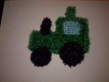 Gehäkelter grüner Traktor aus schimmerndem Effektgarn mit schwarzen Rädern und hellblauem Fenster.