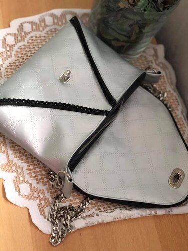 Silberne gesteppte Schultertasche mit schwarzer Zierkante, Drehverschluss und Metallkettenriemen auf Häkeldecke.