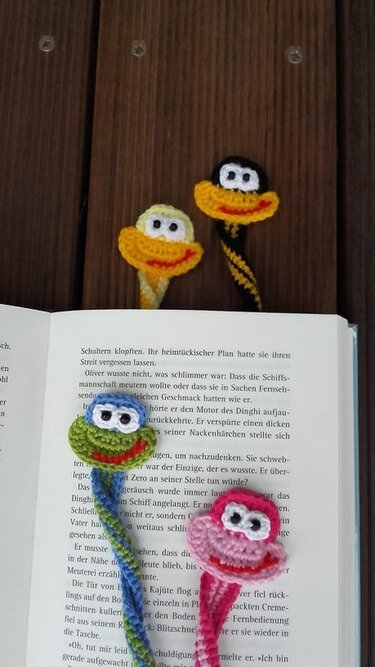 Fünf gehäkelte Tier-Lesezeichen, zwei auf Holz, drei in einem offenen Buch, verschiedene Farben