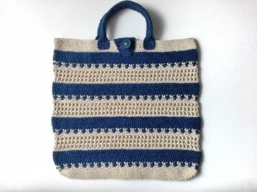 Handgehäkelter Shopper mit horizontalen Streifen in Marineblau und Beige, blauen Tragegriffen und Knopfverschluss auf weißem Hintergrund.