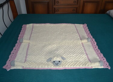 Cremefarbene handgehäkelte Babydecke mit Noppenmuster, rosa Rand und grauer Schäfchen-Applikation auf dunklem Untergrund