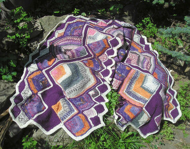 Gestrickter lila Schal mit bunten Quadraten und weißer gezackter Kante auf Felsen und Pflanzen.