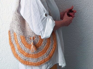 Runde gehäkelte Umhängetasche mit beige-orange Streifen und kleiner Quaste, über der Schulter getragen
