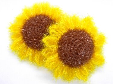 Pair of crocheted sunflower-shaped scrubbers with bright yellow fuzzy petals and brown textured centers on a white background