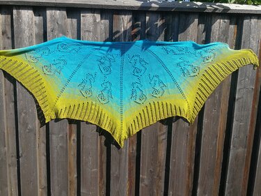 Halbrundes gestricktes Tuch in Türkis und Gelbgrün mit Lochmuster‑Motiven und offener Rippenkante, an einem Holzzaun gespannt