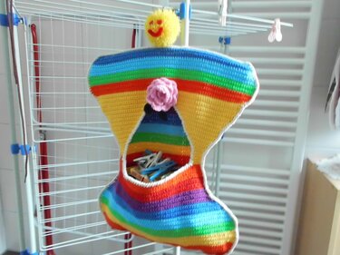 Gehäkelter Wäscheklammerbeutel mit Regenbogenstreifen, rosa Blume und gelbem Smiley, gefüllt mit Wäscheklammern an weißem Wäscheständer.