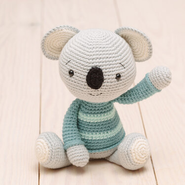 Light grey crocheted koala amigurumi wearing a teal and light‑green striped sweater, arm raised.