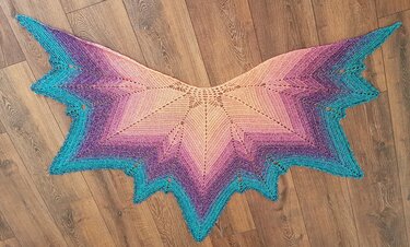 Gehäkelte Stola mit Farbverlauf von Pfirsich über Violett zu Türkis und gezacktem Rand auf Holzboden