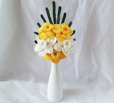 Gehäkelter Narzissenstrauß mit gelben und weißen Blüten und grünen Stielen in einer weißen Vase vor hellem Hintergrund