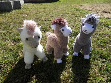 Drei gestrickte Alpakas in Weiß, Beige und Grau mit flauschigen Mähnen