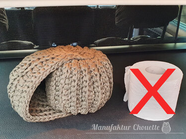 Beigefarbene, grob gehäkelte Hülle neben einer Toilettenpapierrolle mit rotem X