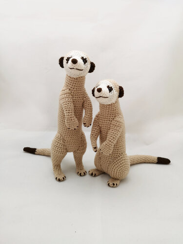 Two crocheted amigurumi meerkats, one standing and one crouching, with light brown bodies, white faces, and dark brown accents.