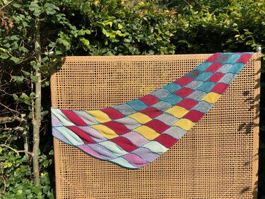 Halbrundes gestricktes Tuch mit Rautenmuster in Rot, Gelb, Türkis, Grau und Lila auf geflochtenem Sichtschutz vor Pflanzen