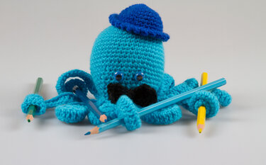 Nahaufnahme gehäkelter türkiser Oktopus mit sichtbarer Maschenstruktur und Tentakeln, die Buntstifte halten