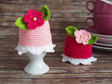Zwei gehäkelte Eierwärmer in Rosa und Rot mit weißer Rüsche, grünen Blättern und Blüten mit Perlen