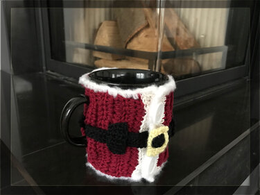 Schwarze Keramiktasse mit rotem gehäkeltem Becherwärmer im Nikolaus-Stil, weißer Kante, schwarzem Gürtel und gelber Schnalle