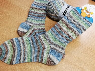 Zwei handgestrickte Socken mit blau‑grün‑braunem Ringelmuster und passendem Wollknäuel auf einem Holztisch.