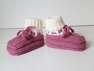 Zwei gestrickte Babyschuhe in Altrosa mit weißem Umschlag auf hellem Hintergrund.