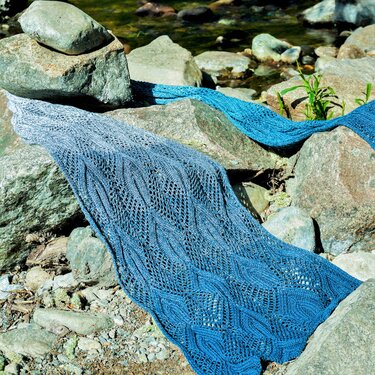 Hand-knitted blue ombre lace shawl draped over river rocks by a stream.
