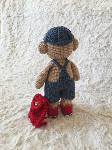 Rückansicht eines gehäkelten Teddybären in blauer Latzhose, passender Mütze, roten Schuhen und kleinem roten Rucksack