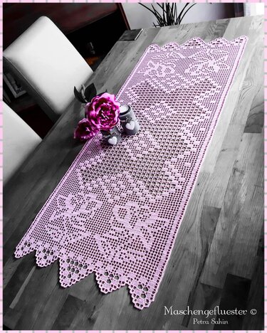 Rosa gehäkelter Filet-Tischläufer mit Blumenmuster und gezacktem Rand auf Holztisch, dekoriert mit Blüten
