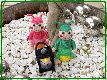 Zwei gehäkelte Käfer-Amigurumi, pink und grün, neben einer schwarzen Häkel-Laterne auf weißen Kieselsteinen