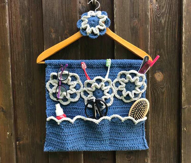 Blauer gehäkelter Wand-Organizer mit Blumenapplikationen und Taschen, aufgehängt an einem Holzkleiderbügel, gefüllt mit Alltagsgegenständen.