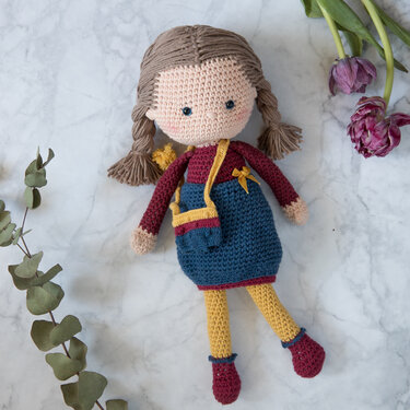 Gehäkelte Amigurumi-Puppe mit braunen Zöpfen, blauem Kleid, gelber Strumpfhose und Umhängetasche auf Marmoruntergrund mit Eukalyptus und Tulpen.