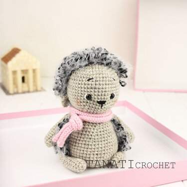 Gehäkelter Amigurumi-Igel mit grauem, flauschigem Rücken und rosa Schal, sitzend