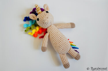 Beiges gehäkeltes Amigurumi‑Einhorn mit goldfarbenem spiralförmigen Horn und regenbogenfarbener lockiger Mähne auf weißem Hintergrund