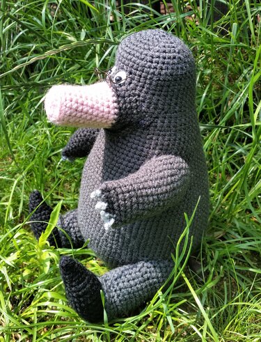 Gehäkelter grauer Amigurumi-Maulwurf mit rosa Schnauze und kleiner Brille sitzt im Gras