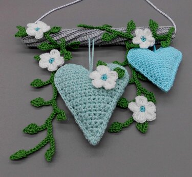 Two light-aqua crocheted hearts hanging from a gray crocheted rod with green crochet vines and white crochet flowers