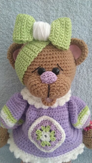 Brauner, gehäkelter Teddybär im lila Kleid mit Granny-Square-Motiv und grüner Schleife vor hellem Hintergrund