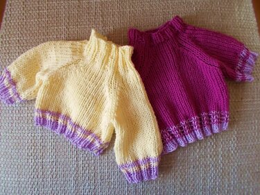 Zwei einfache gestrickte Kinderpullover, gelb und magenta, mit kontrastfarbenen Rippenbündchen, liegend auf hellem Untergrund