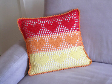 Gehäkeltes Kissen in Rot, Orange und Gelb mit Herzmuster auf Sofa.