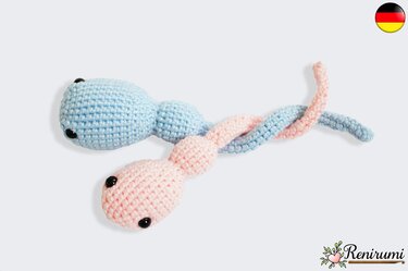 Zwei ineinander verschlungene gehäkelte Amigurumi-Figuren in Hellblau und Zartrosa vor hellem Hintergrund.