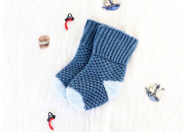 Zweifarbige handgehäkelte Babysocken in dunklem Blau mit hellblauem Zehen- und Fersenbereich auf weißem, flauschigem Untergrund.